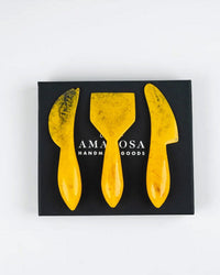 The Casa Amarosa Resin Cheese Knife Set of 3 in Yellow features uniquely shaped handmade knives, displayed on a black box labeled “AMACOSA HANDMADE GOODS”—ideal accessories for any charcuterie board.