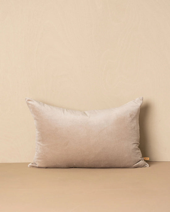 The Saarde Velvet Lumbar Pillow w/ Feather Insert in Stone stands upright against a matching wall and surface, featuring a beige velvet finish and a small fabric tag on the right side.