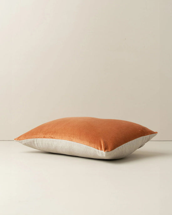 The Saarde Velvet Lumbar Pillow w/ Feather Insert in Terracotta features a burnt orange top and light beige bottom. Resting on a plain, light surface, its feather insert offers extra comfort and support.