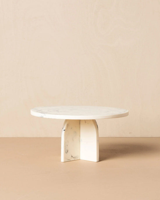 The Saarde Flow Resin Cake Stand in Merle features a round, white marble-look top with subtle grey veining and a thick X-shaped base, shown against a neutral beige background.