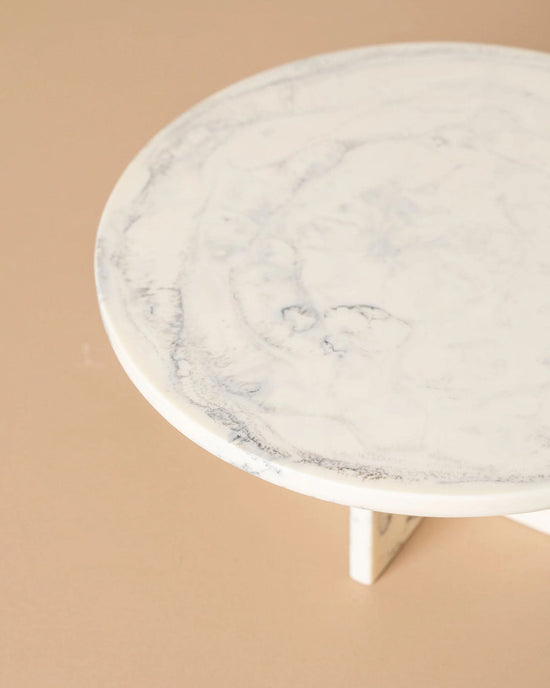 The Flow Resin Cake Stand in Merle by Saarde features a round surface with a handmade marble finish and subtle gray veining, partially shown with one leg against a light beige background.