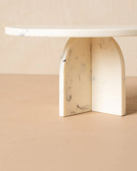 The Flow Resin Cake Stand in Merle by Saarde features a minimalist white marble finish with a round top and two arched legs, displayed on a beige surface against a neutral background.