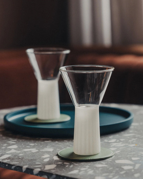 Two Saarde Kairos Wine Glasses in Opaque White, partially filled, rest on green coasters—one sits on a terrazzo table and the other is placed on a blue tray in the background.