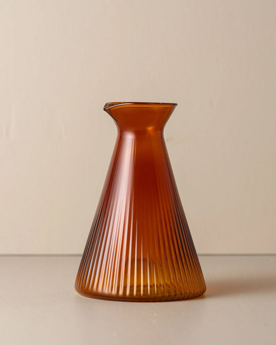 The Kairos Angular Carafe in Amber by Saarde, crafted from borosilicate glass with a flared base and narrow neck, is displayed on a plain surface against a beige background.
