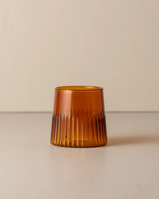 The Saarde Kairos Medium Water Glass in Dark Amber, featuring vertical ridges and a wide shape, sits on a neutral surface with a plain background.