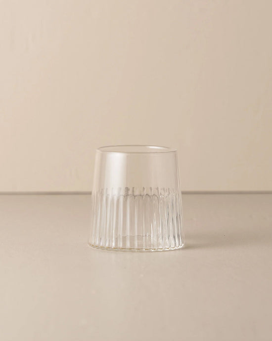 The Kairos Medium Water Glass in Clear by Saarde, made from borosilicate glass with vertical ridges, sits upside down on a smooth neutral surface against a plain beige background.