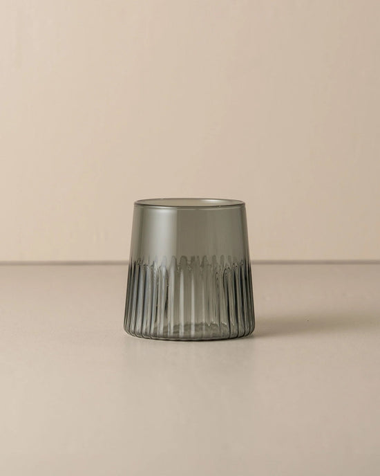 The Saarde Kairos Medium Water Glass in Smoke, featuring a smoky gray tint, is displayed on a beige surface with a matching background.