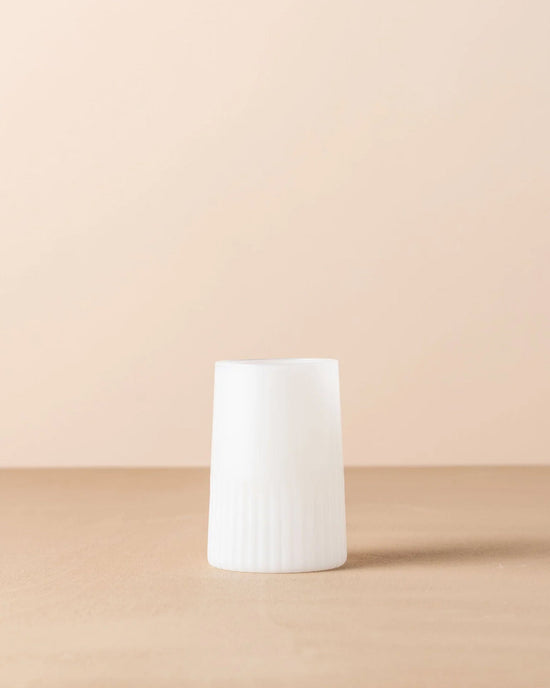 The Saarde Kairos Large Water Glass in Opaque White, featuring a cylindrical shape with a ridged lower half, stands upright on a beige surface against a light beige background.