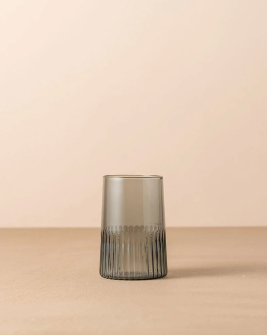 The Saarde Kairos Large Water Glass in Smoke, featuring a textured ribbed base, sits on a plain beige surface against a neutral background.