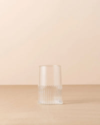 The Saarde Kairos Large Water Glass in Clear, featuring vertical textured lines around the base, is placed on a flat, light brown surface against a beige background.