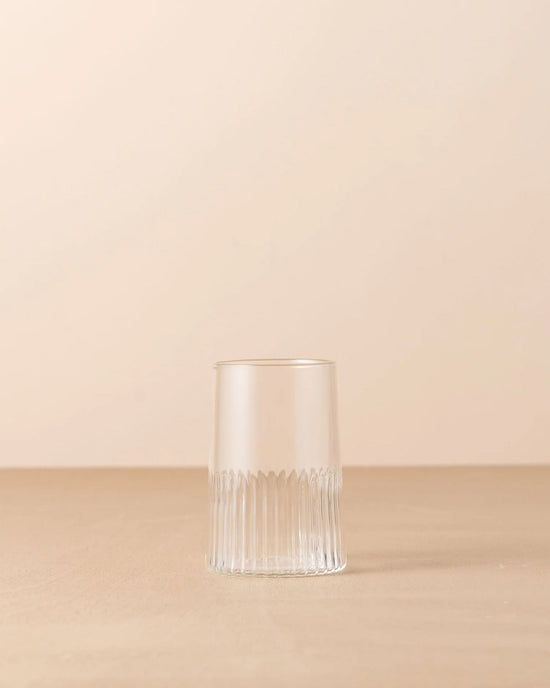 The Saarde Kairos Large Water Glass in Clear, featuring vertical textured lines around the base, is placed on a flat, light brown surface against a beige background.