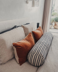 A bed near a window with sheer curtains features two rust pillows, two beige velvet cushions, and a Saarde Velvet Lumbar Pillow w/ Feather Insert in Terracotta.