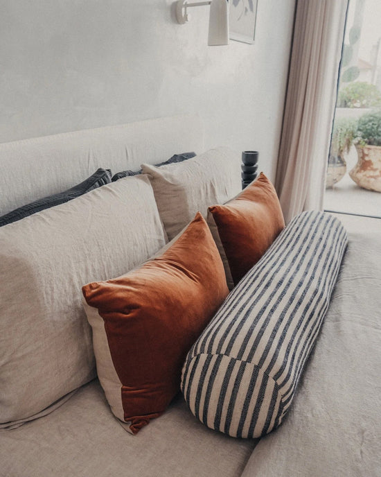 A bed near a window with sheer curtains features two rust pillows, two beige velvet cushions, and a Saarde Velvet Lumbar Pillow w/ Feather Insert in Terracotta.
