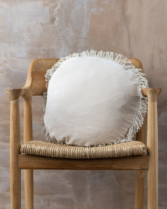A Saarde Velvet Round Pillow w/ Feather Insert in Clay, featuring fringe edging, sits on a wooden chair with a woven seat against a textured beige wall.