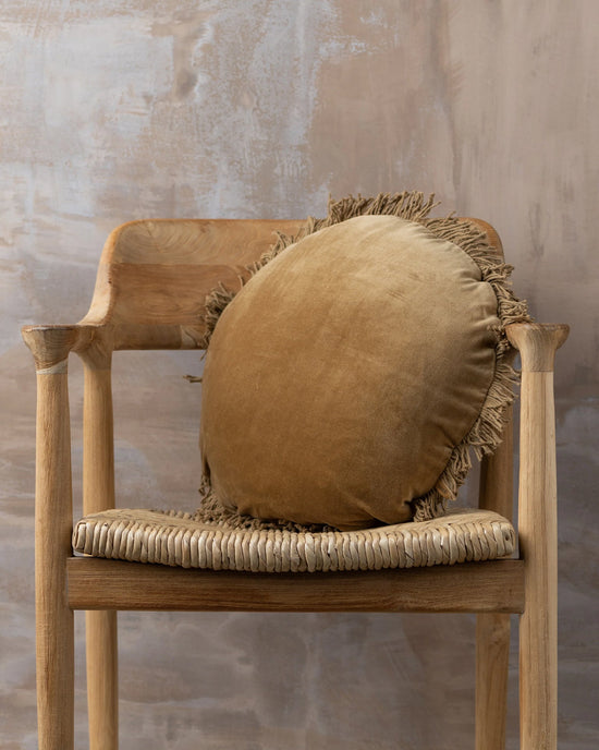 A Saarde Velvet Round Pillow with Feather Insert in Nutmeg rests on a wooden chair with a woven seat, set against a textured beige wall.