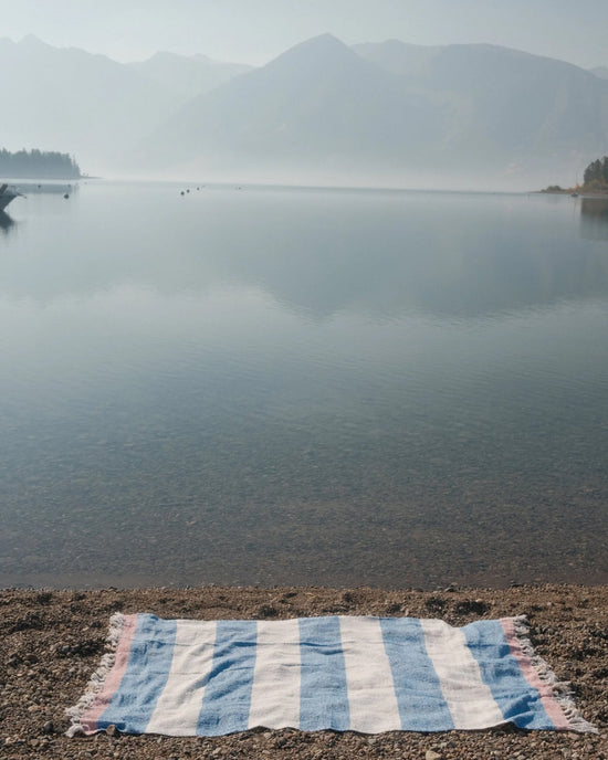 The caminito Suave Aire - Blanket Roll in blue and white stripes rests on a pebbled lakeshore, with misty mountains and trees visible across the calm water.