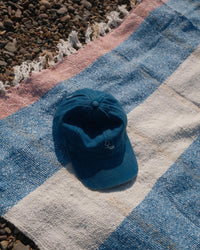 A blue baseball cap rests on a caminito Suave Aire - Blanket Roll—an indoor-outdoor blanket with blue, white, and pink stripes—spread out on a pebble-covered surface.