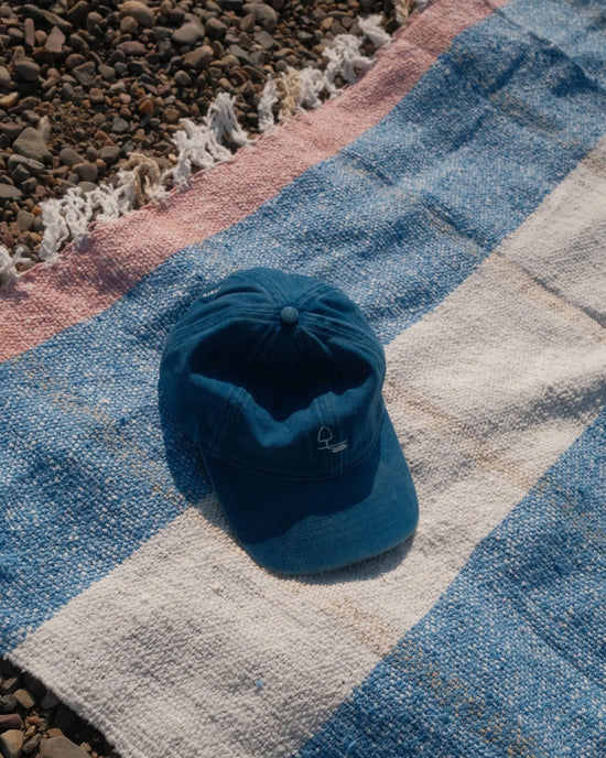 A blue baseball cap rests on a caminito Suave Aire - Blanket Roll—an indoor-outdoor blanket with blue, white, and pink stripes—spread out on a pebble-covered surface.