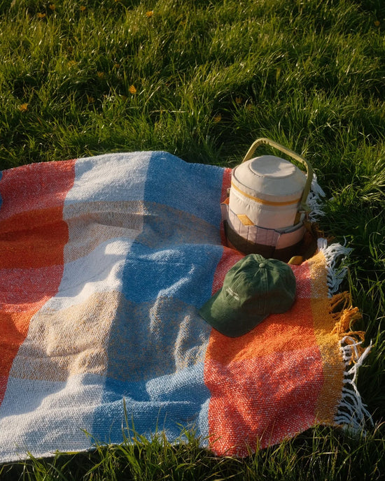 The Suave Paseo - Blanket Roll by caminito, a colorful handwoven throw with a leather carrying strap, is spread on green grass alongside a green baseball cap and thermos jug—perfect for an outdoor picnic or indoor lounging.