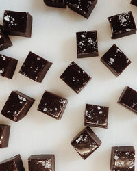 Wildflower Caramel Co’s Dark Chocolate Sea Salt Caramels in a Bag are glossy squares sprinkled with coarse sea salt, artfully arranged on a white surface for an irresistible presentation.