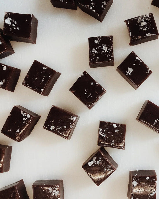 Wildflower Caramel Co’s Dark Chocolate Sea Salt Caramels in a Bag are glossy squares sprinkled with coarse sea salt, artfully arranged on a white surface for an irresistible presentation.