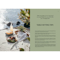 A lakeside picnic with bread, cheese, fruit, and juice captures the spirit of "Four Seasons of Cabin Cooking" by Cozy Publishing, as sunlight sparkles on the water and greenery frames the rocks—Nordic-inspired table tips appear alongside.