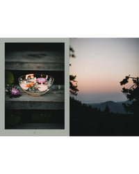 The "Nordic Magic" bowl by Cozy Publishing, featuring floating flowers and candles inspired by nature worship, rests on a wood surface beside a scenic sunset over forested hills under a colorful sky.
