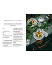 A page from the Nordic Summer Cookbook by Cozy Publishing appears beside two plates of rösti with herbs, cream, and salmon—ideal for a Scandinavian-inspired midsummer table set outdoors with vintage dishes and flowers.
