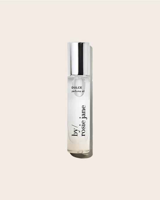 A clear glass bottle of Rosie Jane Perfume Oil in Dulce with a silver cap stands upright against a plain light beige background, highlighting this elegant roll-on fragrance.