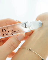 A hand applies Rosie Jane Perfume Oil in Dulce to a wrist with a delicate chain bracelet, highlighting the clear vanilla allure of this roll-on fragrance.