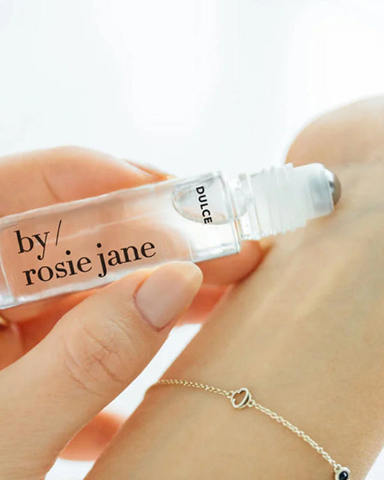 A hand applies Rosie Jane Perfume Oil in Dulce to a wrist with a delicate chain bracelet, highlighting the clear vanilla allure of this roll-on fragrance.