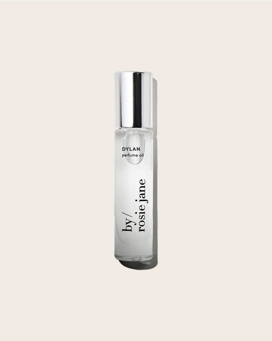 A clear glass bottle of Rosie Jane Perfume Oil in Dylan with a silver cap sits on a light beige background, showcasing this unisex fragrance’s effortless sophistication.