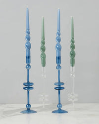 Four spiral candles, two blue and two green, are displayed in matching etúHOME Clear Glass Candlesticks Set of 2 - Medium on a white surface against a gray background.