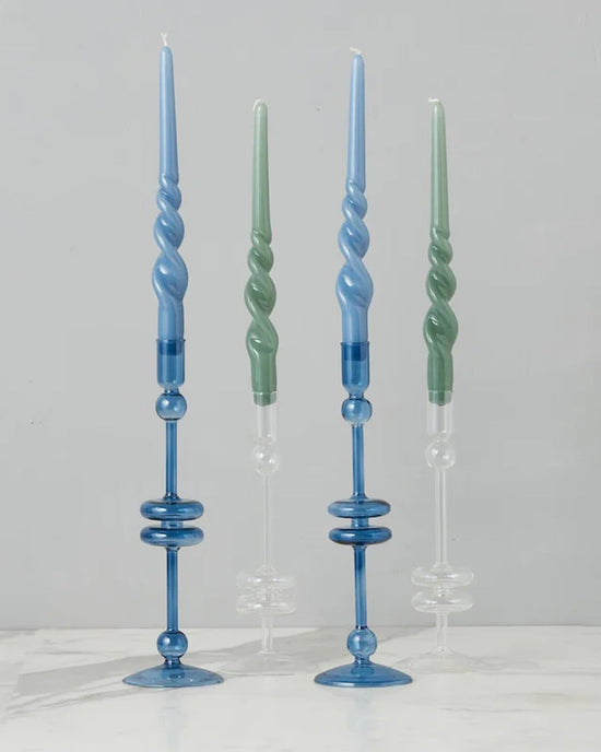 Four spiral candles, two blue and two green, are displayed in matching etúHOME Clear Glass Candlesticks Set of 2 - Medium on a white surface against a gray background.