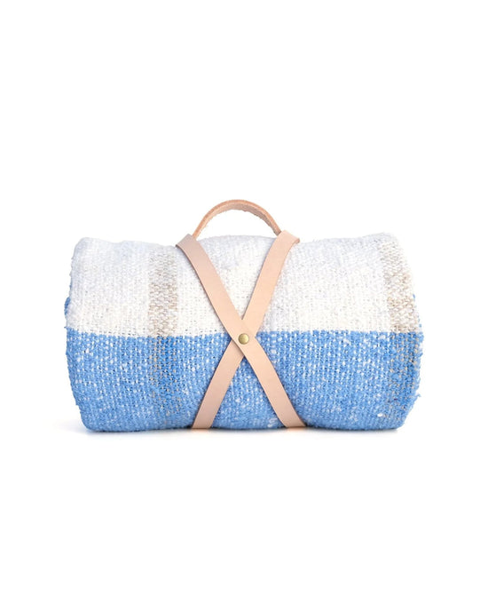 The Suave Aire - Blanket Roll by caminito is a handwoven indoor/outdoor blanket in blue, white, and beige, secured with a tan leather strap and handy handle. Displayed against a white background.