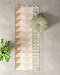 A geometric runner rug with tan and green triangle patterns rests on a light gray floor. The Casa Amarosa Velvet Round Pillow in Pistachio Green adds a decorative touch on one side, while a leafy green plant is placed nearby.