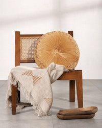 A wooden chair with a woven backrest holds a beige geometric throw and the Casa Amarosa Velvet Round Pillow in Clay. Two wooden trays rest nearby, completing this cozy, minimalist setting.