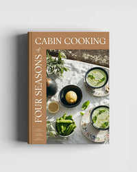 Four Seasons of Cabin Cooking by Cozy Publishing features a cover with bowls of green soup, lemon, snap peas, and herbs on rustic wood—evoking the spirit of outdoor and seasonal cabin recipes.