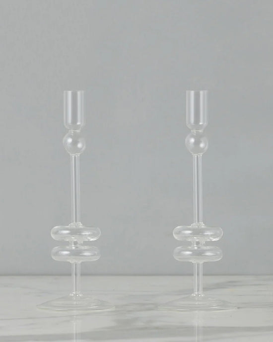 The etúHOME Clear Glass Candlesticks Set of 2 - Medium features handmade recycled glass holders with rounded accents, shown on a white marble surface against a plain gray background.