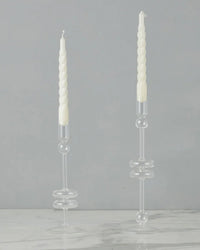 Two clear glass candlesticks (Set of 2, Medium) by etúHOME hold white spiral taper candles on a white marble surface, set against a plain gray background.