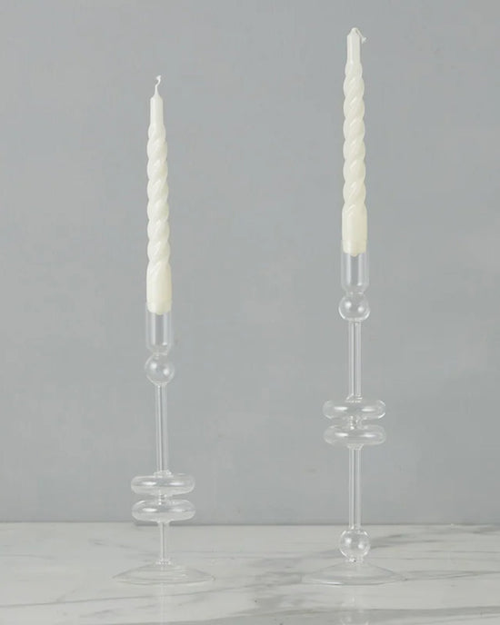 Two clear glass candlesticks (Set of 2, Medium) by etúHOME hold white spiral taper candles on a white marble surface, set against a plain gray background.