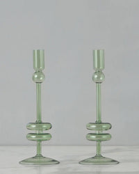 Two medium Sage Glass Candlesticks from etúHOME, crafted by hand from recycled glass with rounded bases, are displayed side by side on a white marble surface against a plain gray background.
