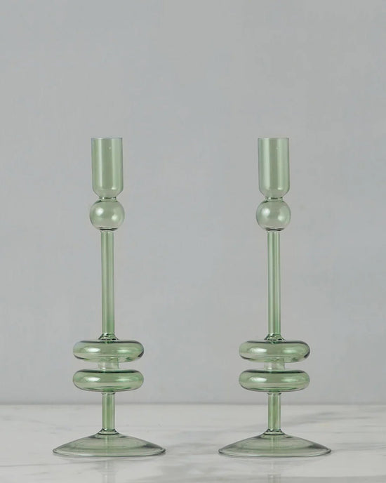 Two medium Sage Glass Candlesticks from etúHOME, crafted by hand from recycled glass with rounded bases, are displayed side by side on a white marble surface against a plain gray background.