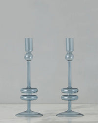 The etúHOME Denim Glass Candlesticks Set of 2 - Medium features modern blue glass holders with circular accents, displayed on white marble against a gray backdrop—an elegant addition to any space for taper candles.