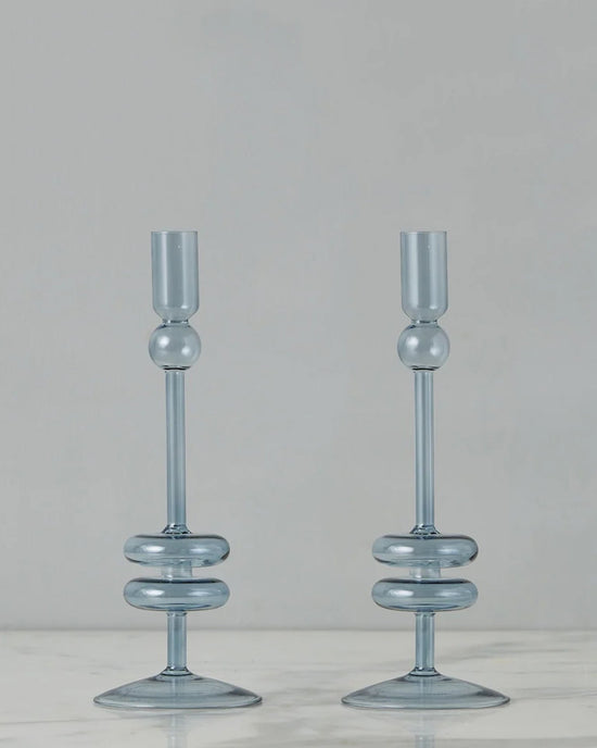 The etúHOME Denim Glass Candlesticks Set of 2 - Medium features modern blue glass holders with circular accents, displayed on white marble against a gray backdrop—an elegant addition to any space for taper candles.