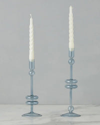 The etúHOME Denim Glass Candlesticks Set of 2 - Medium, crafted from recycled glass, display white spiral taper candles elegantly on a white marble surface against a plain gray background.