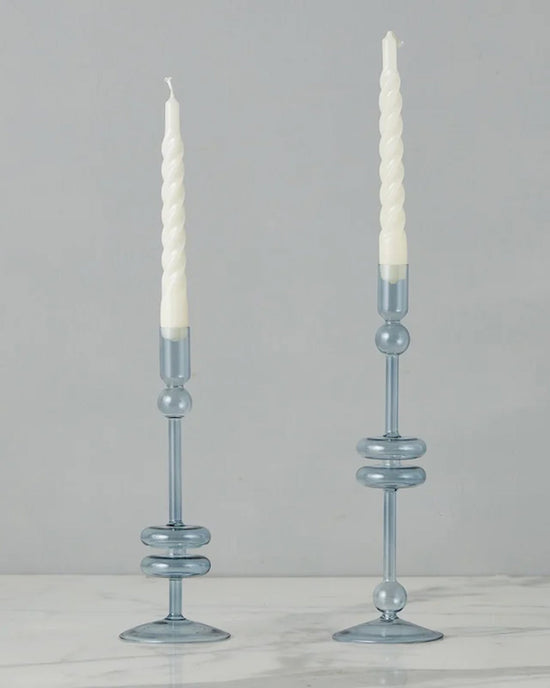 The etúHOME Denim Glass Candlesticks Set of 2 - Medium, crafted from recycled glass, display white spiral taper candles elegantly on a white marble surface against a plain gray background.