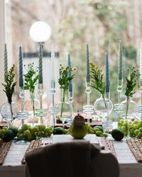 A dining table set with etúHOME Sage Glass Candlesticks (Set of 2, Medium), pale blue candles, green grapes, limes, and greenery. Place settings feature a pear labeled Gabby. The candlesticks are handmade from recycled glass.