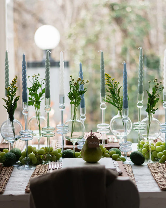 A dining table set with etúHOME Sage Glass Candlesticks (Set of 2, Medium), pale blue candles, green grapes, limes, and greenery. Place settings feature a pear labeled Gabby. The candlesticks are handmade from recycled glass.