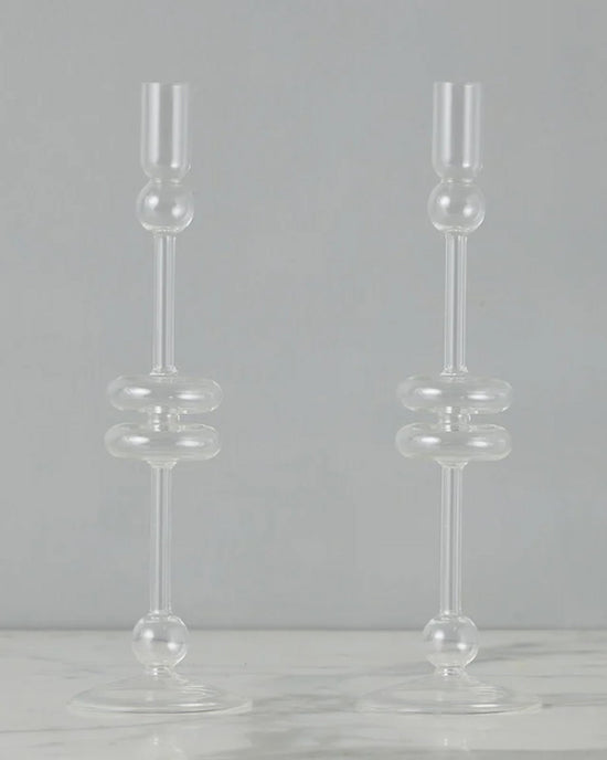 The etúHOME Clear Glass Candlesticks Set of 2 - Large features modern, geometric round and cylindrical shapes, displayed on a light surface with a gray background.
